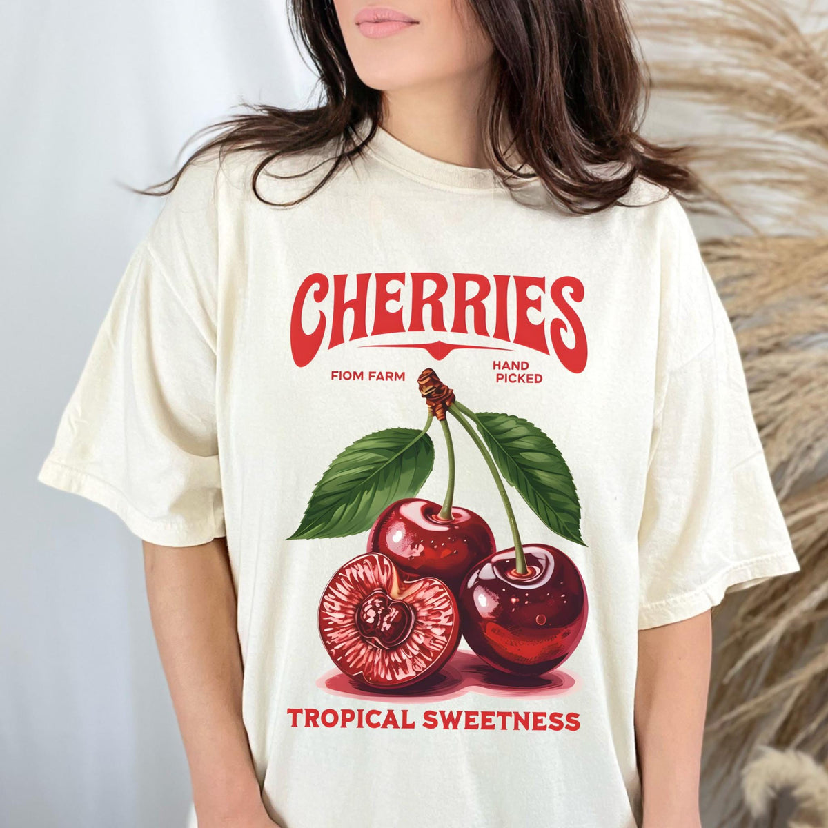 May include: Cream-colored t-shirt featuring a vintage-style cherry graphic. The design includes the word "CHERRIES" in red, along with "FIOM FARM" and "HAND PICKED." Below the cherries, the text reads "TROPICAL SWEETNESS."