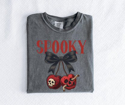 May include: A gray t-shirt with the word "SPOOKY" in red block letters above a black bow and two red cherries. The cherries have skull faces and worms. The shirt is folded on a white surface.
