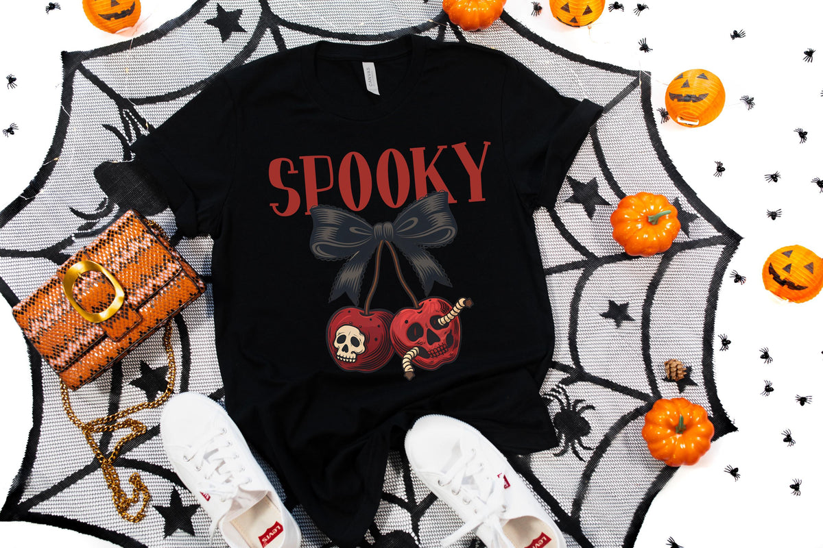 May include: Black t-shirt with the word "SPOOKY" in red, featuring a bow and cherry design with skull details. The image includes a woven orange purse, white sneakers, and Halloween pumpkins.