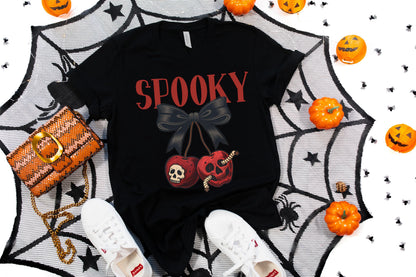 May include: Black t-shirt with the word "SPOOKY" in red, featuring a bow and cherry design with skull details. The image includes a woven orange purse, white sneakers, and Halloween pumpkins.