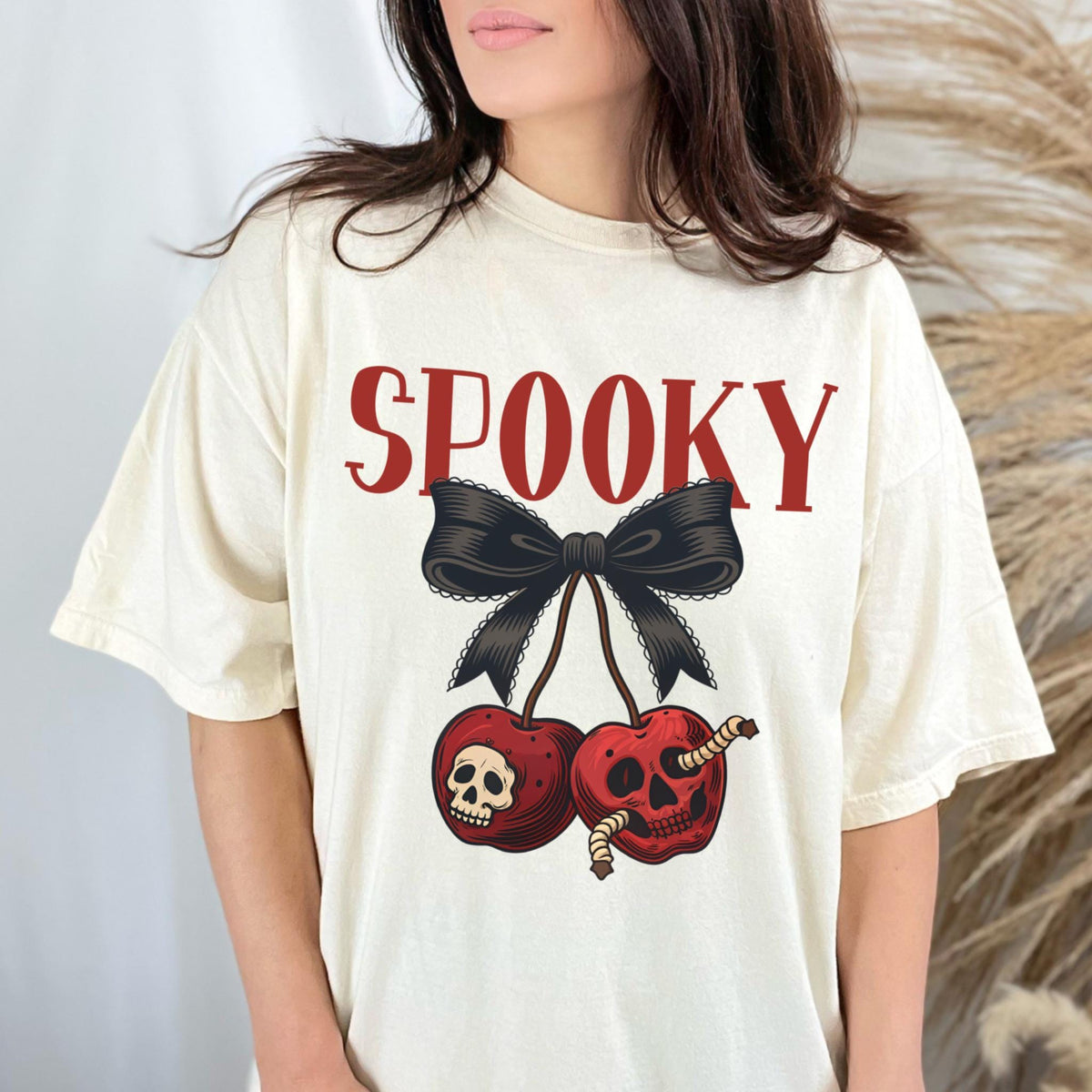 May include: Cream-colored t-shirt with the word "SPOOKY" in red block letters above a black bow and two red cherries. The cherries have skull and worm graphics, creating a Halloween theme.