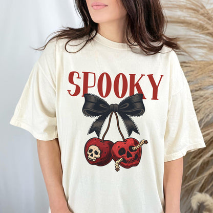 May include: Cream-colored t-shirt with the word "SPOOKY" in red block letters above a black bow and two red cherries. The cherries have skull and worm graphics, creating a Halloween theme.