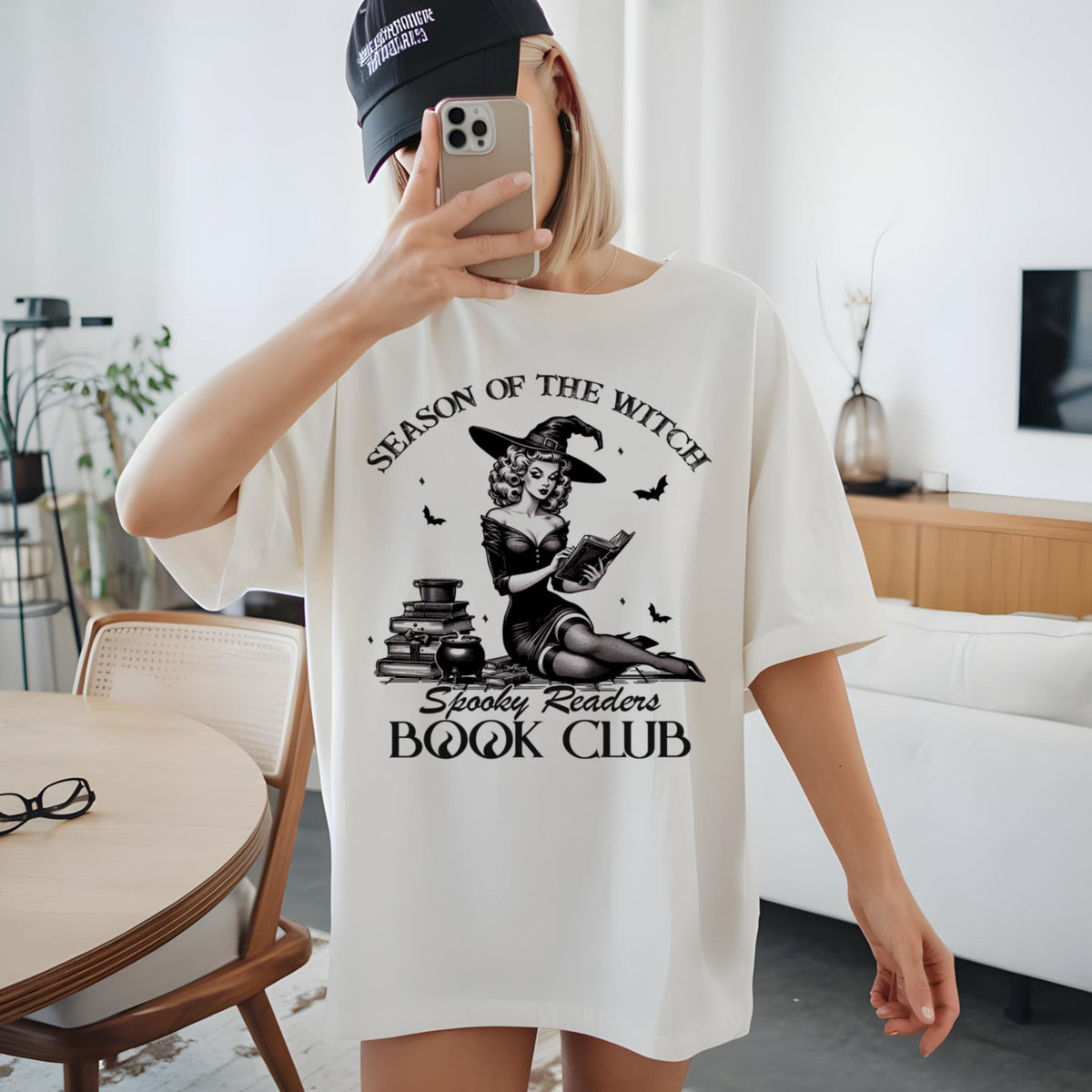 May include: Cream-colored t-shirt featuring a black and white illustration of a witch reading a book, with the text "Season of the Witch Spooky Readers Book Club". The design includes bats and stacks of books.