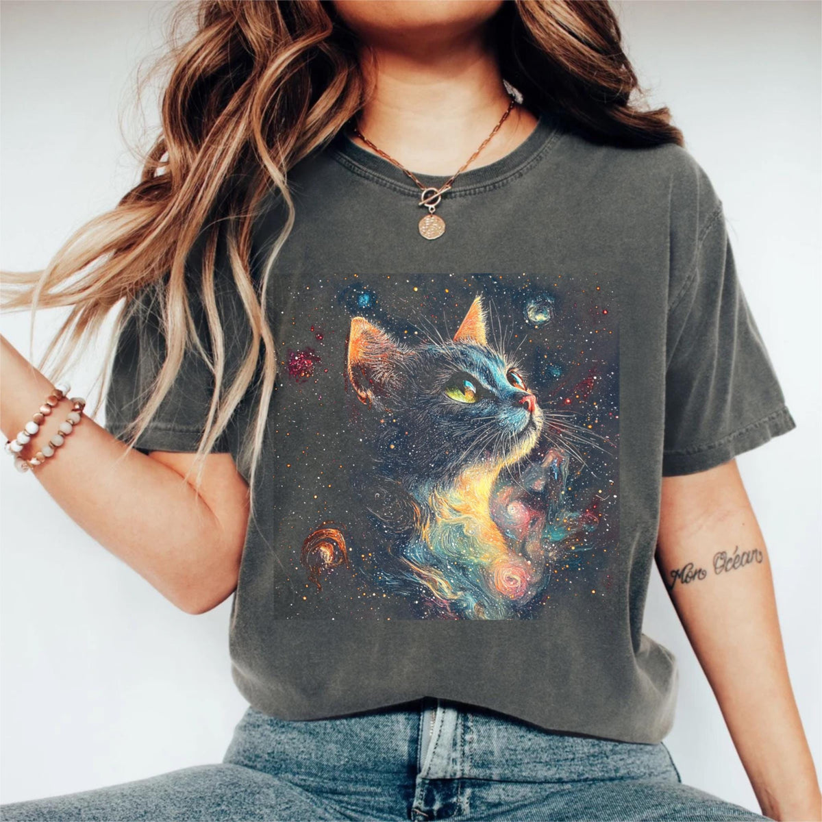 May include: Dark gray t-shirt featuring a vibrant graphic of a cat with a galaxy design. The cat has green eyes and is looking upwards. The artwork includes colorful nebulae and stars, creating a cosmic theme.
