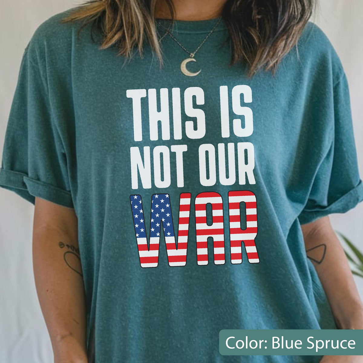 May include: Blue spruce t-shirt with the text "THIS IS NOT OUR WAR" in white. The word "WAR" features a design of the American flag. The shirt is a solid color and has rolled-up sleeves.