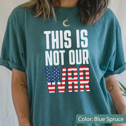 May include: Blue spruce t-shirt with the text "THIS IS NOT OUR WAR" in white. The word "WAR" features a design of the American flag. The shirt is a solid color and has rolled-up sleeves.