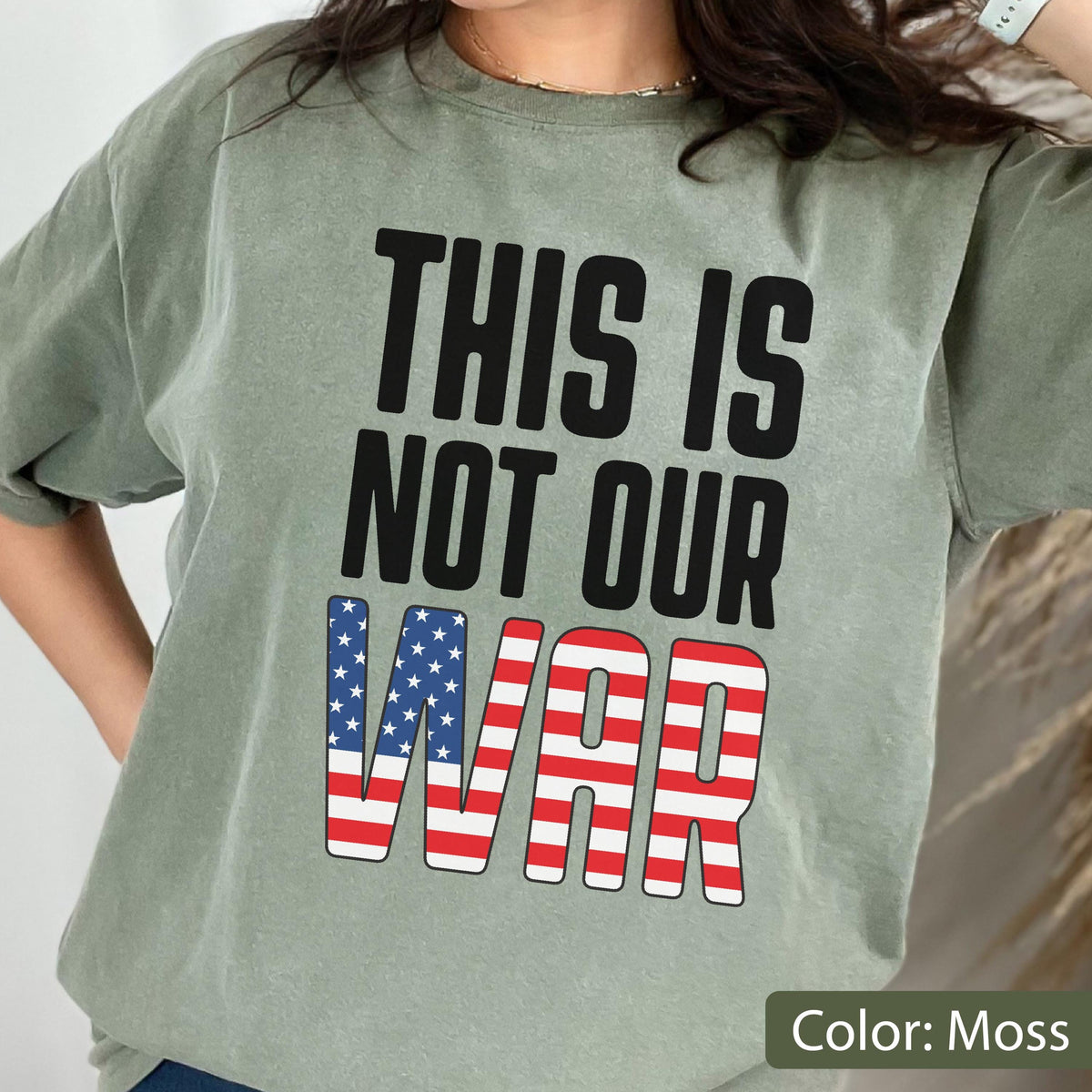 May include: A moss green t-shirt with the text "THIS IS NOT OUR WAR" in black block letters. The word "WAR" is designed with the American flag's stars and stripes. The shirt is a casual style.