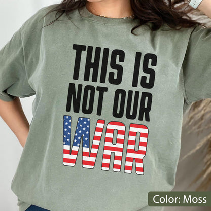May include: A moss green t-shirt with the text "THIS IS NOT OUR WAR" in black block letters. The word "WAR" is designed with the American flag's stars and stripes. The shirt is a casual style.