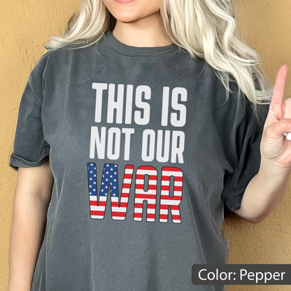 May include: Pepper-colored t-shirt with the text "THIS IS NOT OUR WAR" in white block letters. The word "WAR" is designed with a red and white striped pattern and blue stars.