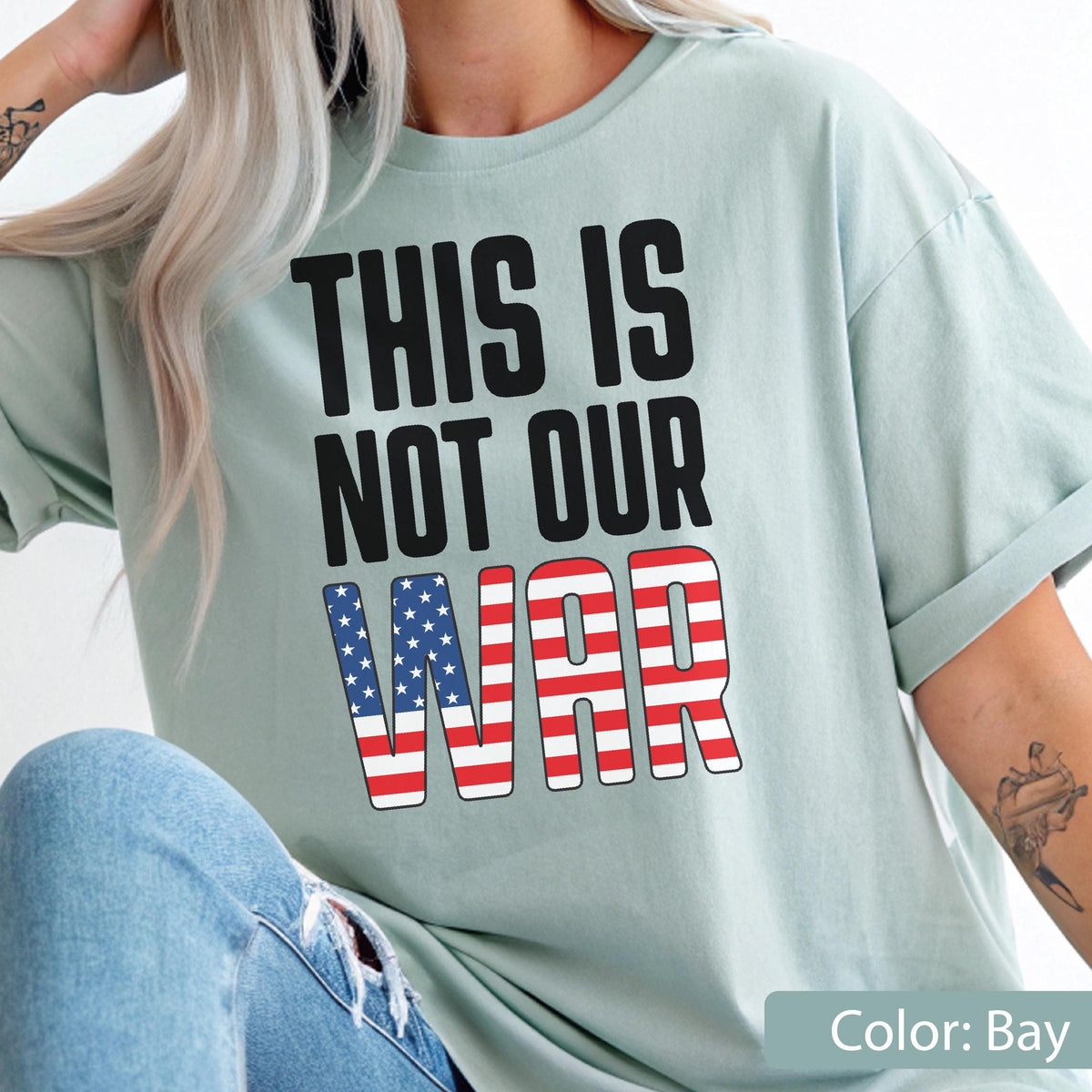 May include: A light blue t-shirt with the text "THIS IS NOT OUR WAR" in black block letters. The word "WAR" is designed with a red, white, and blue American flag pattern. The shirt is a solid color and has short sleeves.