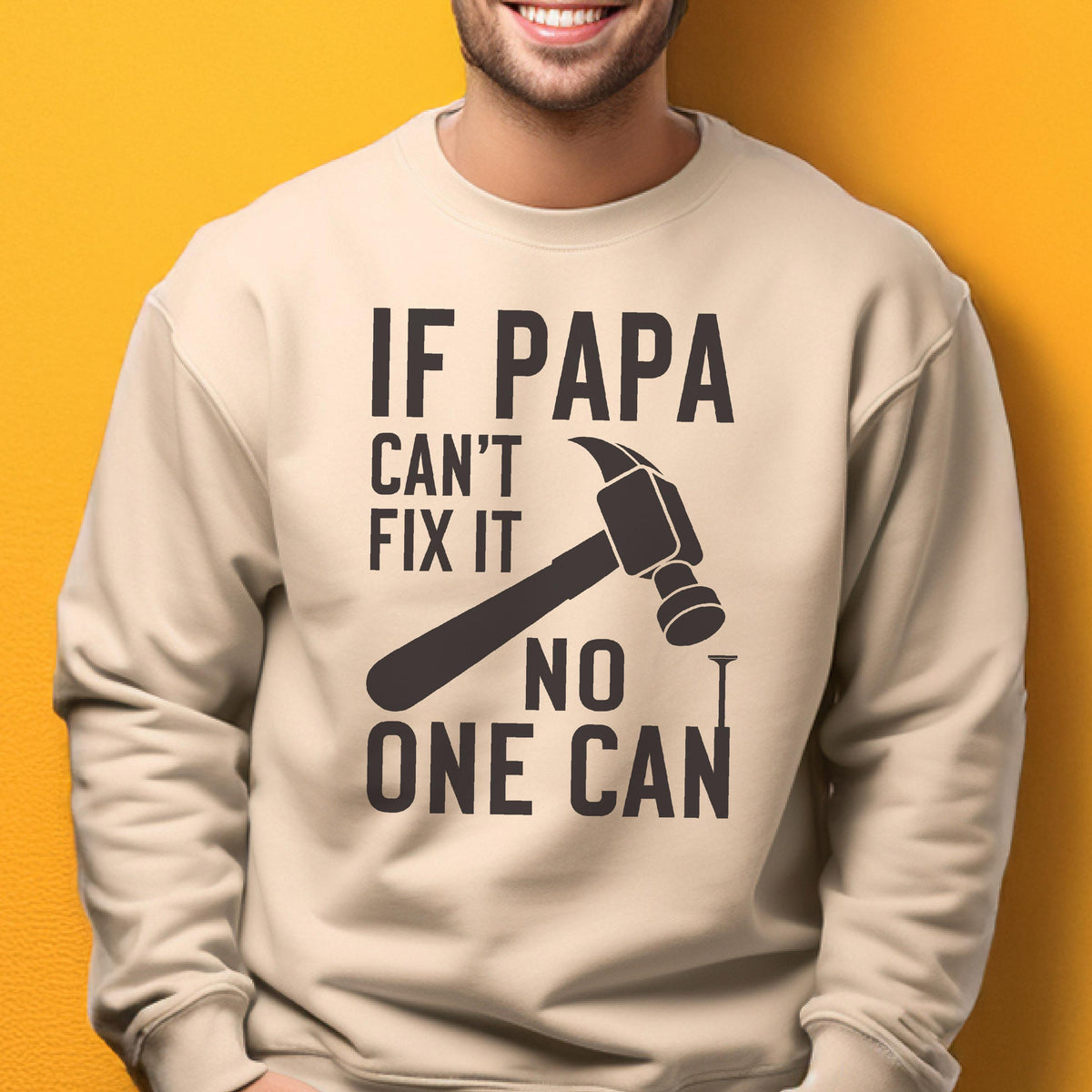 May include: A beige sweatshirt with the text "IF PAPA CAN'T FIX IT NO ONE CAN" in dark brown. A hammer and nail graphic are featured in the center of the design. The sweatshirt is a casual style.
