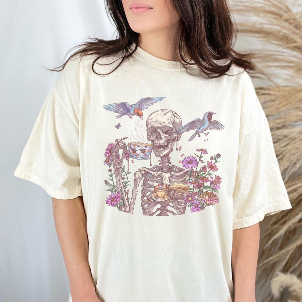 May include: Cream-colored t-shirt featuring a whimsical illustration of a skeleton enjoying tea amidst flowers and birds. The skeleton holds a teacup, with steam rising. The design includes colorful flowers and two birds in flight.