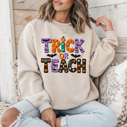 May include: Beige sweatshirt with a colorful Halloween-themed graphic. The design features the words "TRICK OR TEACH" in orange, purple, and green, along with bats, pumpkins, and spiderwebs. The sweatshirt is a casual, long-sleeved garment.
