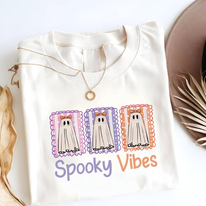 May include: Cream-colored t-shirt featuring a graphic of three cartoon ghosts with bows, set in decorative frames. The text "Spooky Vibes" is below the ghosts. A gold necklace is draped on the shirt.