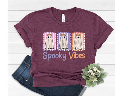May include: A maroon t-shirt featuring a graphic of three cartoon ghosts in postage stamp frames. Each ghost has a bow and is set against a different pastel background. The words "Spooky Vibes" are printed below the ghosts.