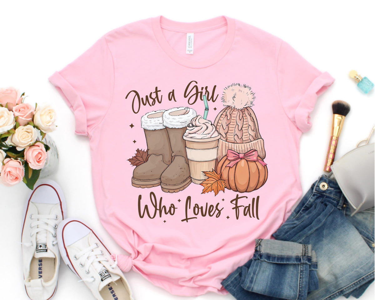 May include: Pink t-shirt with the text "Just a Girl Who Loves Fall." The graphic includes brown boots, a coffee drink, a knit hat, and a pumpkin. White sneakers and a bouquet of flowers are also visible.