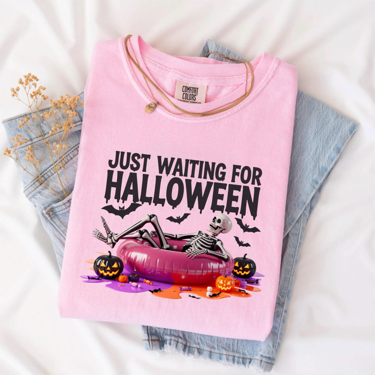 May include: A light pink t-shirt with the text "JUST WAITING FOR HALLOWEEN" and a skeleton relaxing in a pink inflatable ring. The design includes black bats and jack-o'-lanterns. A gold necklace and folded blue jeans are also visible.