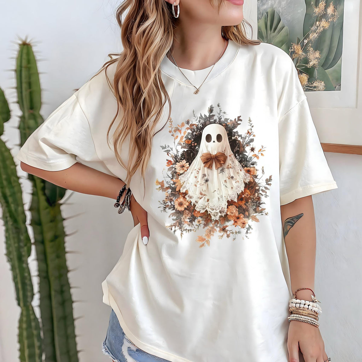 May include: Cream-colored t-shirt featuring a whimsical ghost design. The ghost is adorned with a bow tie and a floral arrangement. The design incorporates shades of brown, orange, and white, creating a vintage aesthetic.