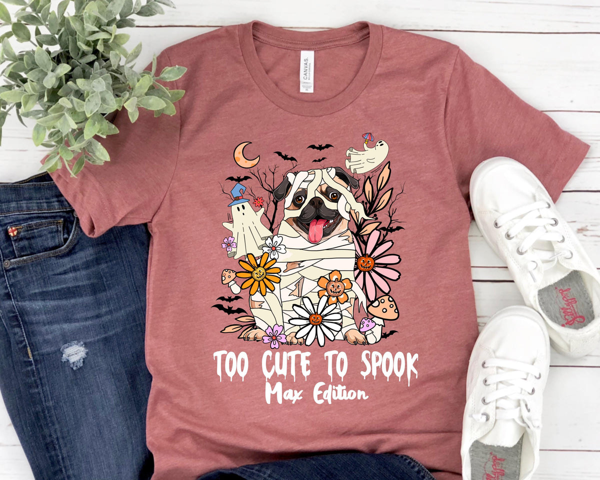 May include: A mauve t-shirt featuring a Halloween-themed graphic. The design includes a pug dressed as a mummy, surrounded by flowers, bats, and a ghost. The text "TOO CUTE TO SPOOK Max Edition" is printed below the graphic.