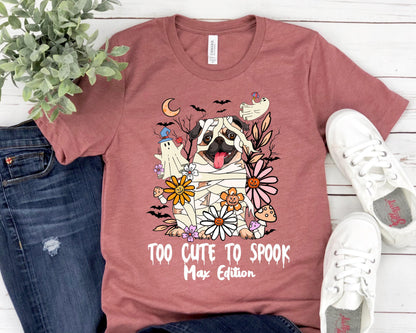 May include: A mauve t-shirt featuring a Halloween-themed graphic. The design includes a pug dressed as a mummy, surrounded by flowers, bats, and a ghost. The text "TOO CUTE TO SPOOK Max Edition" is printed below the graphic.