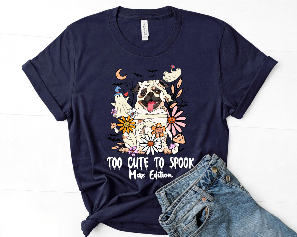 May include: Navy blue t-shirt featuring a cartoon pug dressed as a mummy, surrounded by flowers, a ghost, and a mushroom. The text "TOO CUTE TO SPOOK Max Edition" is printed below the graphic. The shirt is paired with blue jeans.