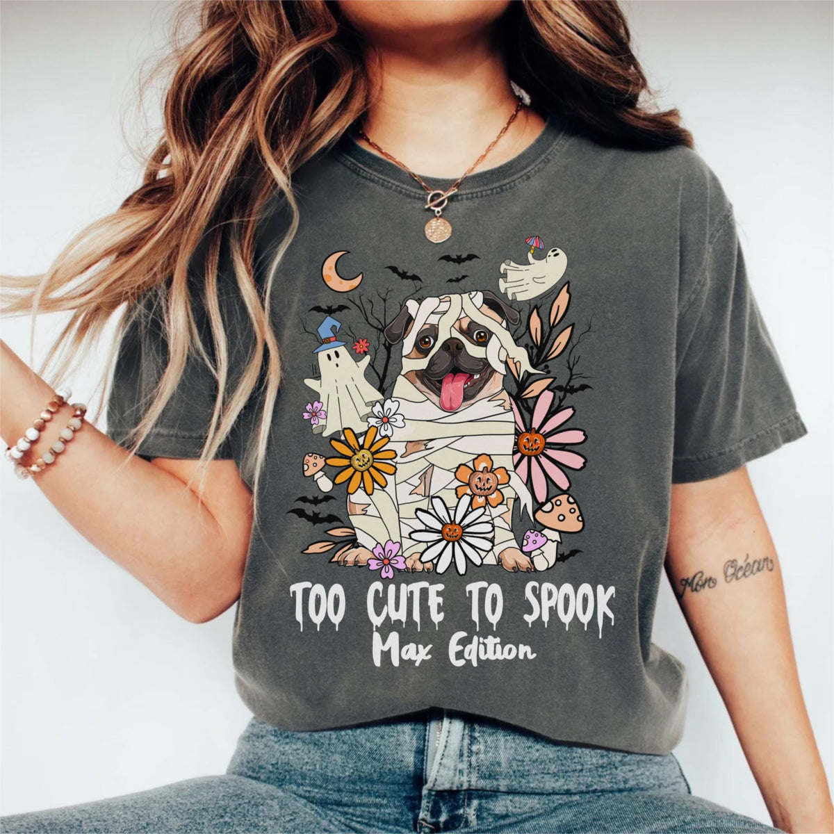 May include: Dark gray t-shirt featuring a cartoon pug dressed as a mummy, surrounded by flowers and ghosts. The text "TOO CUTE TO SPOOK Max Edition" is printed below the graphic. The shirt has a crew neck and short sleeves.