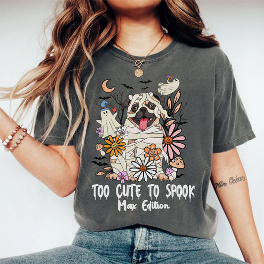 May include: Dark gray t-shirt featuring a cartoon pug dressed as a mummy, surrounded by flowers and ghosts. The text "TOO CUTE TO SPOOK Max Edition" is printed below the graphic. The shirt has a crew neck and short sleeves.