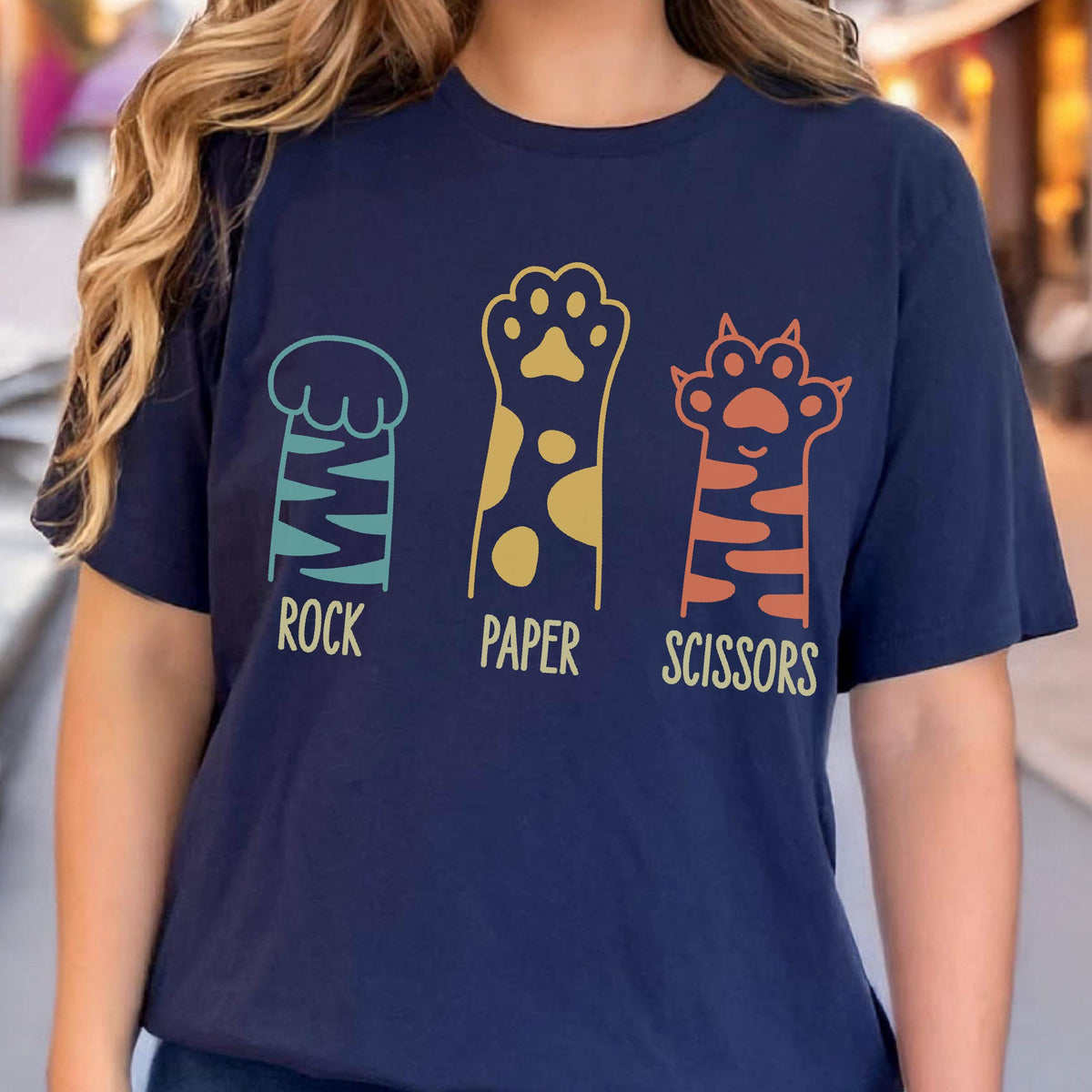 May include: A navy blue t-shirt featuring a graphic of three animal paws representing the game rock, paper, scissors. The first paw is teal with stripes, the second is gold with spots, and the third is orange with stripes, each labeled below.
