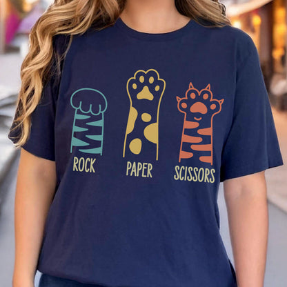 May include: A navy blue t-shirt featuring a graphic of three animal paws representing the game rock, paper, scissors. The first paw is teal with stripes, the second is gold with spots, and the third is orange with stripes, each labeled below.