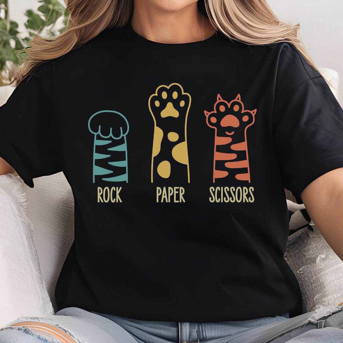 May include: A black t-shirt featuring a graphic of three animal paws representing the game Rock Paper Scissors. The first paw is teal with stripes, the second is yellow with spots, and the third is orange with stripes. The words ROCK, PAPER, SCISSORS are printed below each respective paw in a light yellow font.