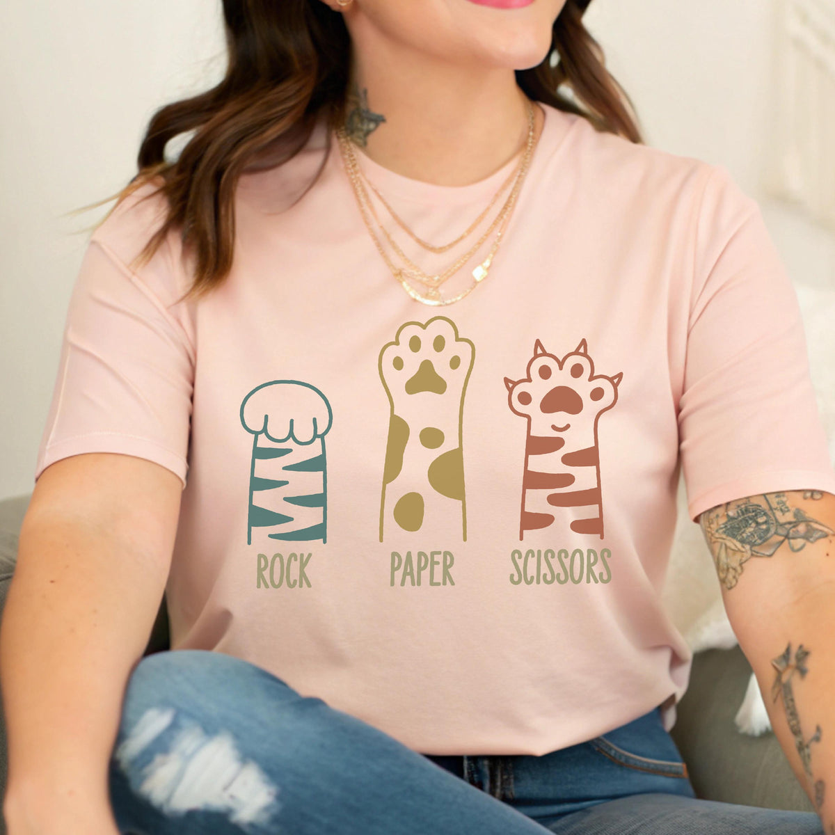 May include: A light pink short-sleeve t-shirt featuring a graphic of three animal paws representing the game Rock, Paper, Scissors. The first paw is teal with stripes, the second is mustard yellow with spots, and the third is terracotta with stripes.