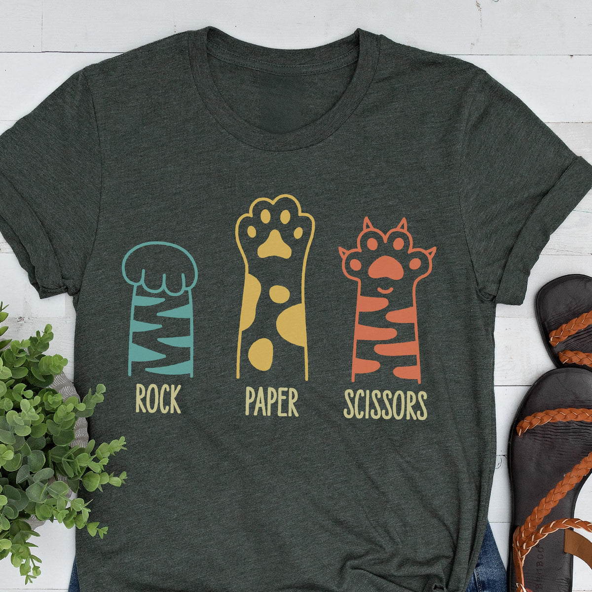 May include: A dark charcoal gray short-sleeve t-shirt featuring a graphic of three animal paws. The first paw is teal with stripes, the second is yellow with spots, and the third is orange with stripes. Below each paw is the text Rock, Paper, Scissors.