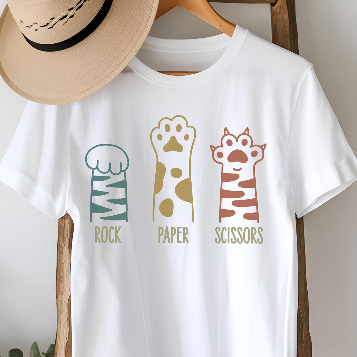 May include: A white short-sleeved t-shirt featuring a graphic of three cat paws with the text ROCK, PAPER, and SCISSORS printed below each paw. The first paw is teal with stripes, the second is gold with spots, and the third is terracotta with stripes.
