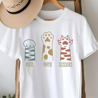 May include: A white short-sleeved t-shirt featuring a graphic of three cat paws with the text ROCK, PAPER, and SCISSORS printed below each paw. The first paw is teal with stripes, the second is gold with spots, and the third is terracotta with stripes.
