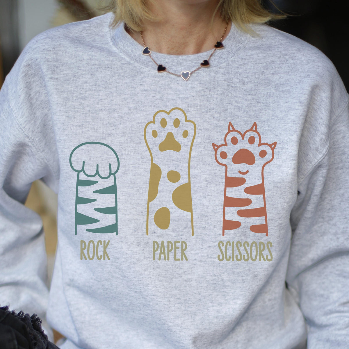 May include: A light gray crewneck sweatshirt featuring a graphic of three animal paws. The first is teal with stripes, the second is gold with spots, and the third is terracotta with stripes. Below each paw is the text Rock, Paper, and Scissors.