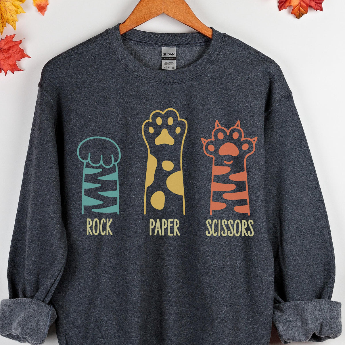 May include: A dark charcoal gray crewneck sweatshirt featuring a graphic of three animal paws representing rock, paper, and scissors. The first paw is teal with stripes, the second is yellow with spots, and the third is orange with stripes.