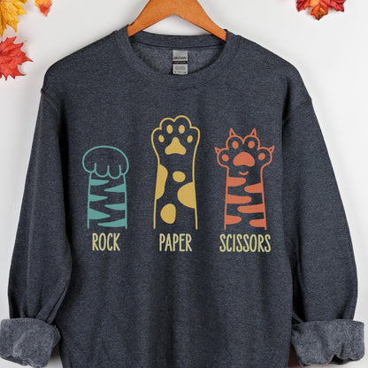 May include: A dark charcoal gray crewneck sweatshirt featuring a graphic of three animal paws representing rock, paper, and scissors. The first paw is teal with stripes, the second is yellow with spots, and the third is orange with stripes.