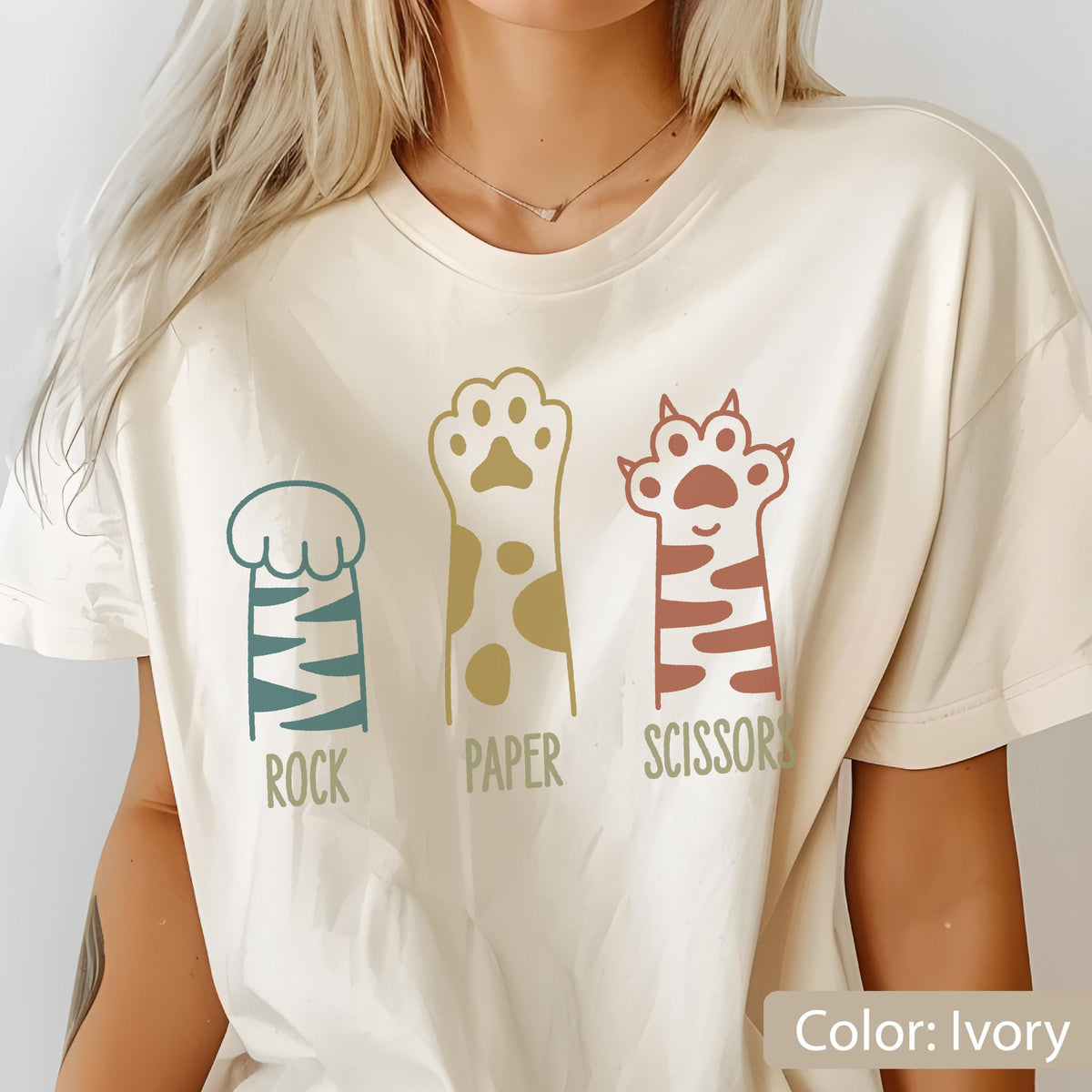 May include: An ivory t-shirt featuring a graphic print of three animal paws arranged side by side. The first paw is teal with stripes, labeled ROCK. The second is gold with spots, labeled PAPER. The third is rust-colored with stripes, labeled SCISSORS.