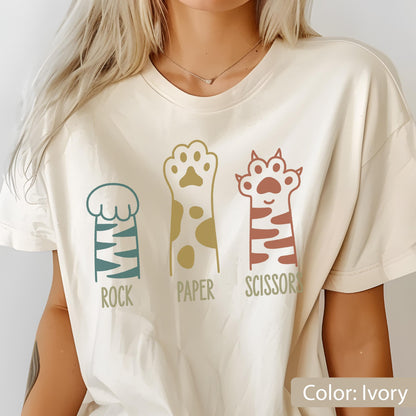 May include: An ivory t-shirt featuring a graphic print of three animal paws arranged side by side. The first paw is teal with stripes, labeled ROCK. The second is gold with spots, labeled PAPER. The third is rust-colored with stripes, labeled SCISSORS.