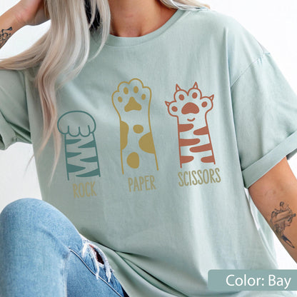 May include: A bay-colored t-shirt featuring a graphic of three animal paws representing the game rock, paper, scissors. The first paw is teal with stripes, the second is gold with spots, and the third is rust-colored with stripes and text.
