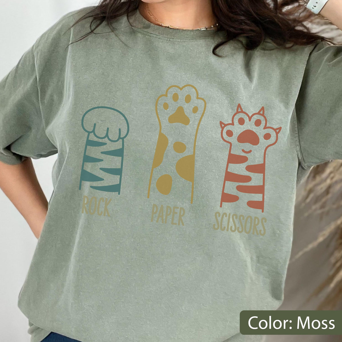 May include: A moss green short-sleeve t-shirt featuring a graphic of three animal paws representing rock, paper, and scissors. The first paw is teal with stripes, the second is mustard yellow with spots, and the third is terracotta with stripes.
