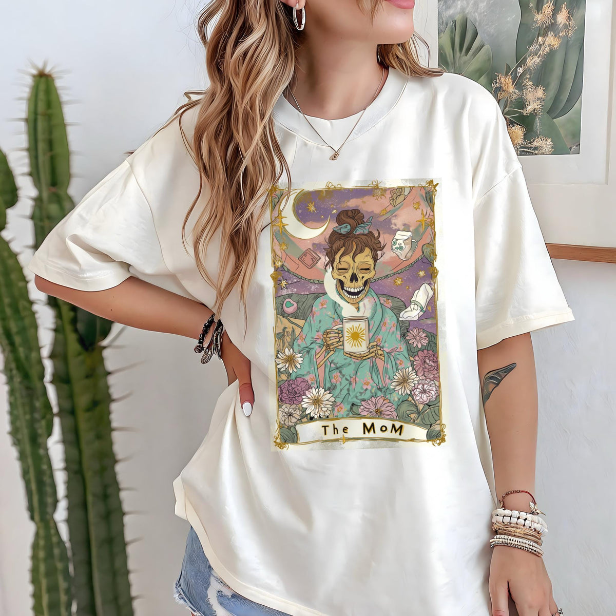 May include: Cream-colored t-shirt featuring a tarot card-style graphic. The graphic depicts a skeleton figure holding a cup, surrounded by flowers and celestial elements. The text "The Mom" is at the bottom of the graphic.