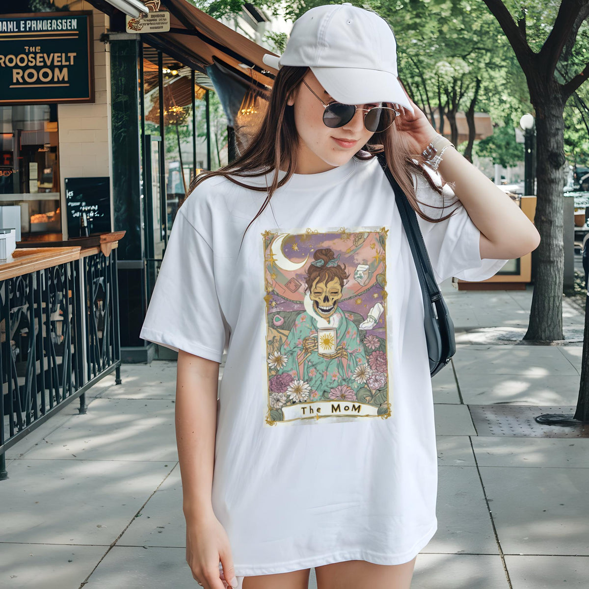 May include: White t-shirt featuring a tarot card design with a skeleton figure holding a sun. The card is framed with floral and celestial elements, with the text "The MoM" at the bottom. The shirt is worn by a person.