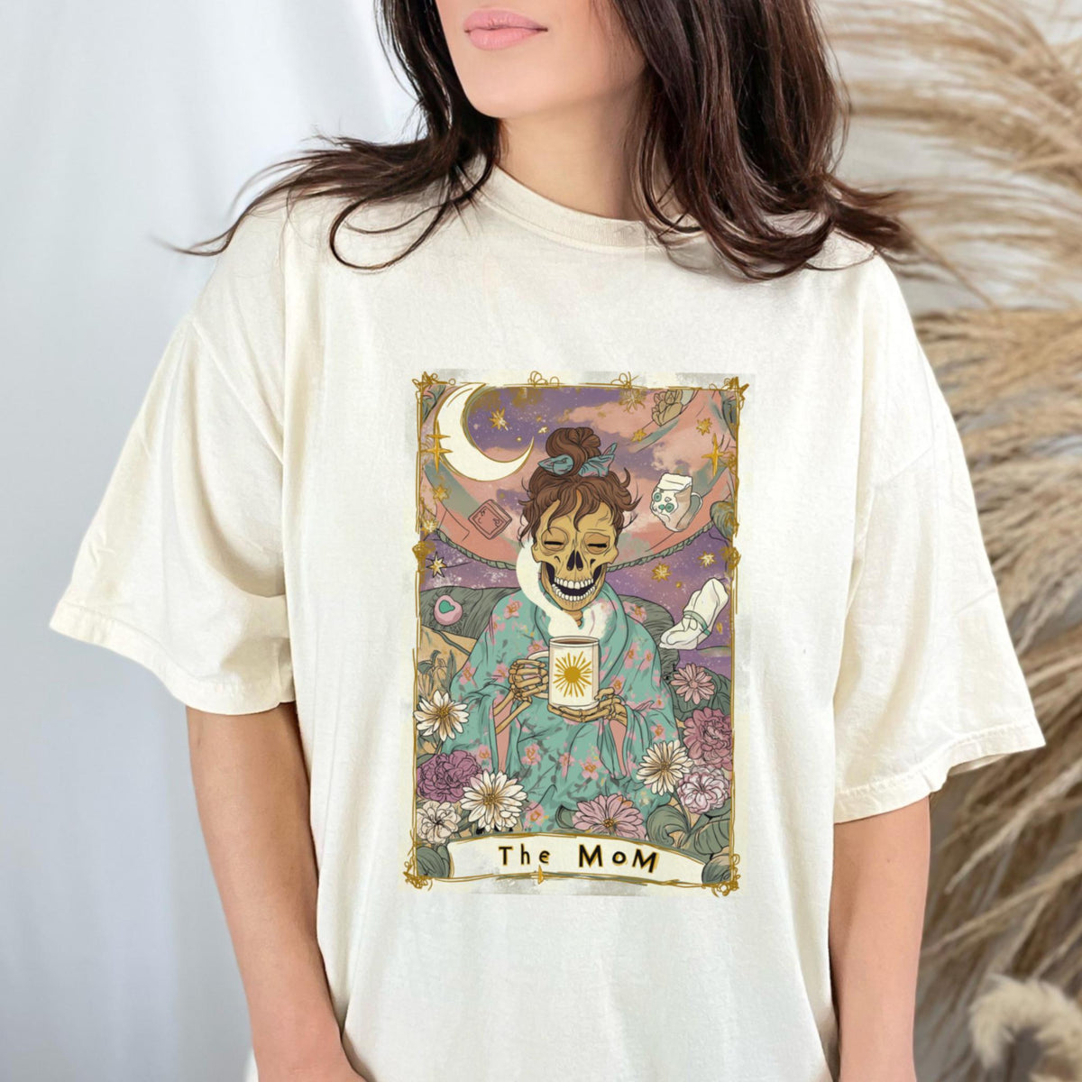 May include: Cream-colored t-shirt featuring a tarot card design with a skeleton figure holding a mug with a sun design. The card is framed with floral and celestial elements, and the text "The MoM" is at the bottom.