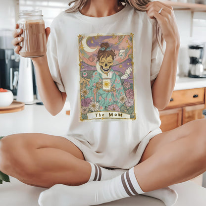 May include: Cream-colored t-shirt featuring a tarot card design with a skeleton figure holding a sun. The card is framed with gold accents and the text "The MoM" is at the bottom. The person is wearing white socks with brown stripes.