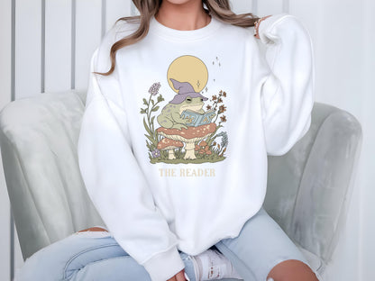 May include: White sweatshirt featuring a whimsical illustration of a frog wearing a wizard hat and reading a book while sitting on a mushroom. The design includes a yellow moon and the text "THE READER".