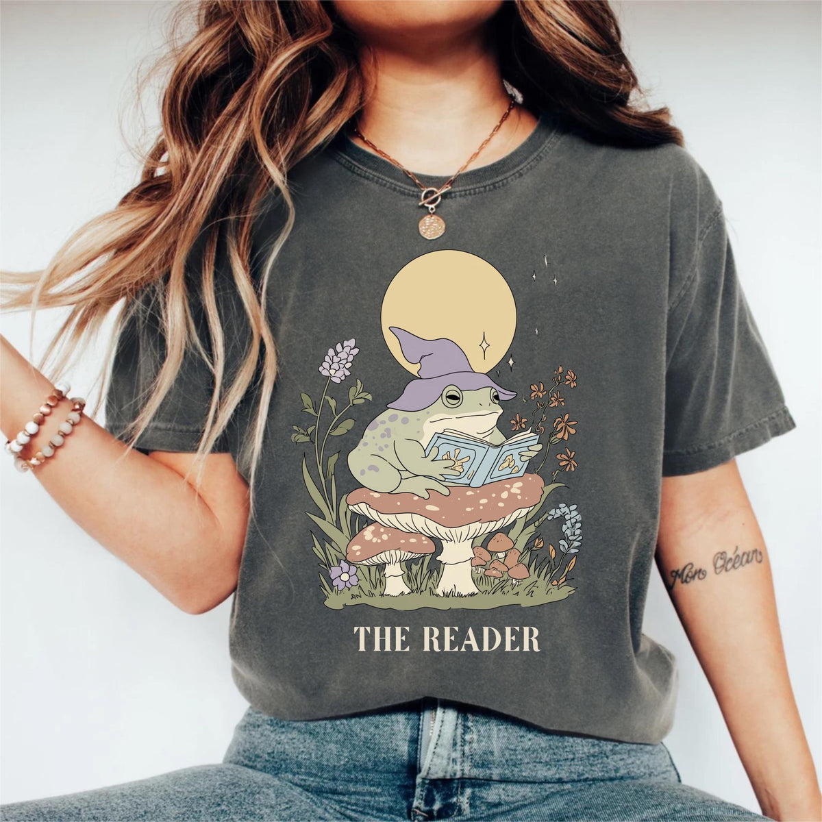 May include: Dark grey t-shirt featuring a whimsical illustration of a frog wearing a wizard hat and reading a book while sitting on a mushroom. The text "THE READER" is printed below the illustration. The design includes a full moon and floral elements.