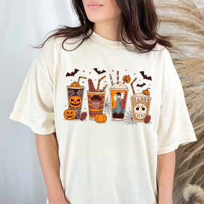 May include: Cream-colored t-shirt featuring a Halloween-themed design. The graphic showcases four stylized coffee cups with spooky characters, pumpkins, bats, and spiderwebs. The cups have straws and are topped with whipped cream. The overall design is festive and fun.