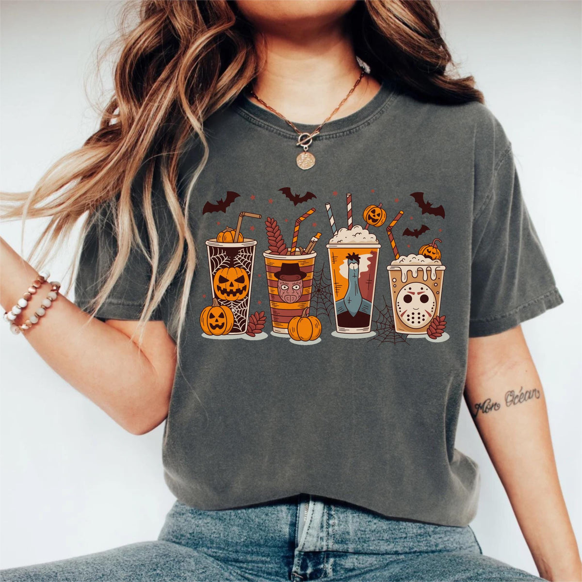 May include: Dark grey t-shirt featuring a Halloween-themed graphic. The design includes five stylized coffee cups with spooky faces, pumpkins, bats, and spiderwebs. The cups have different designs, including a jack-o'-lantern, a character with a hat, and a hockey mask.