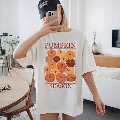 May include: Cream-colored t-shirt featuring a graphic of pumpkins in shades of orange and brown, with the words "PUMPKIN SEASON" in a brown font. The shirt has a relaxed fit and short sleeves.